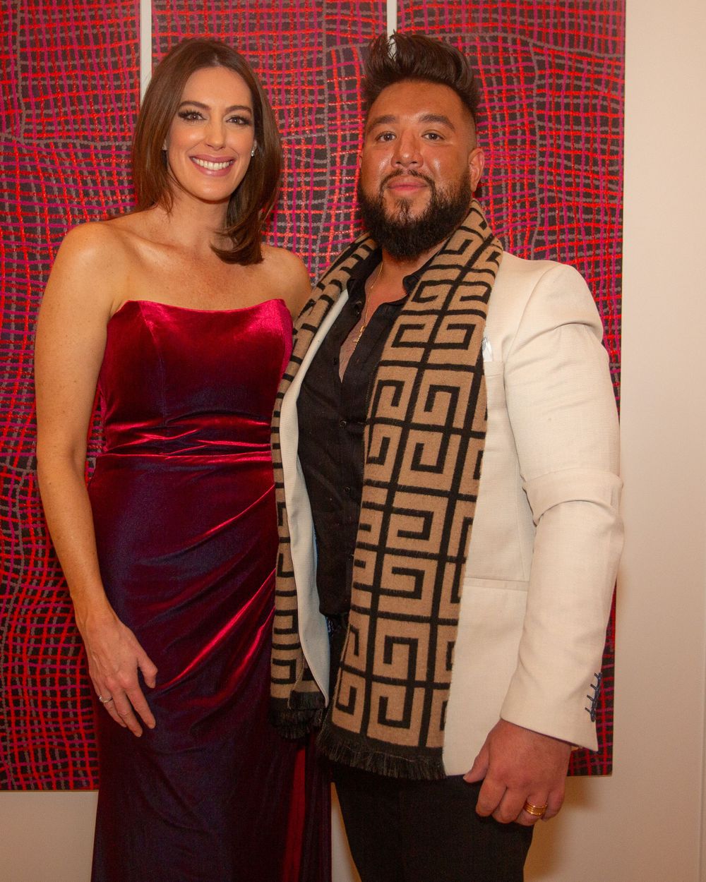 A couple poses together against a red patterned backdrop wearing formal attire.