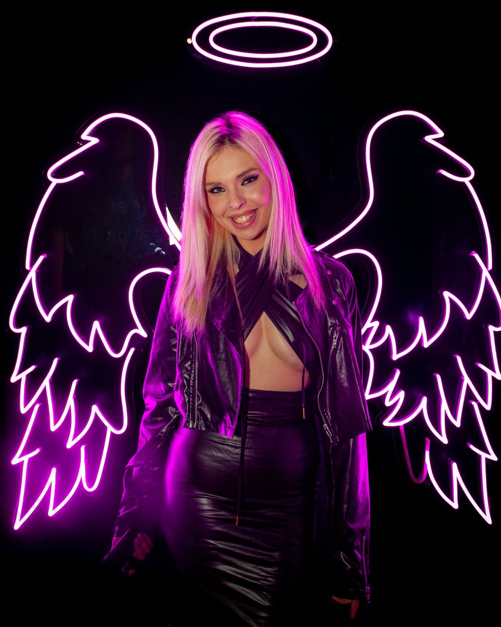 Woman poses against neon angel wings backdrop in purple leather jacket.