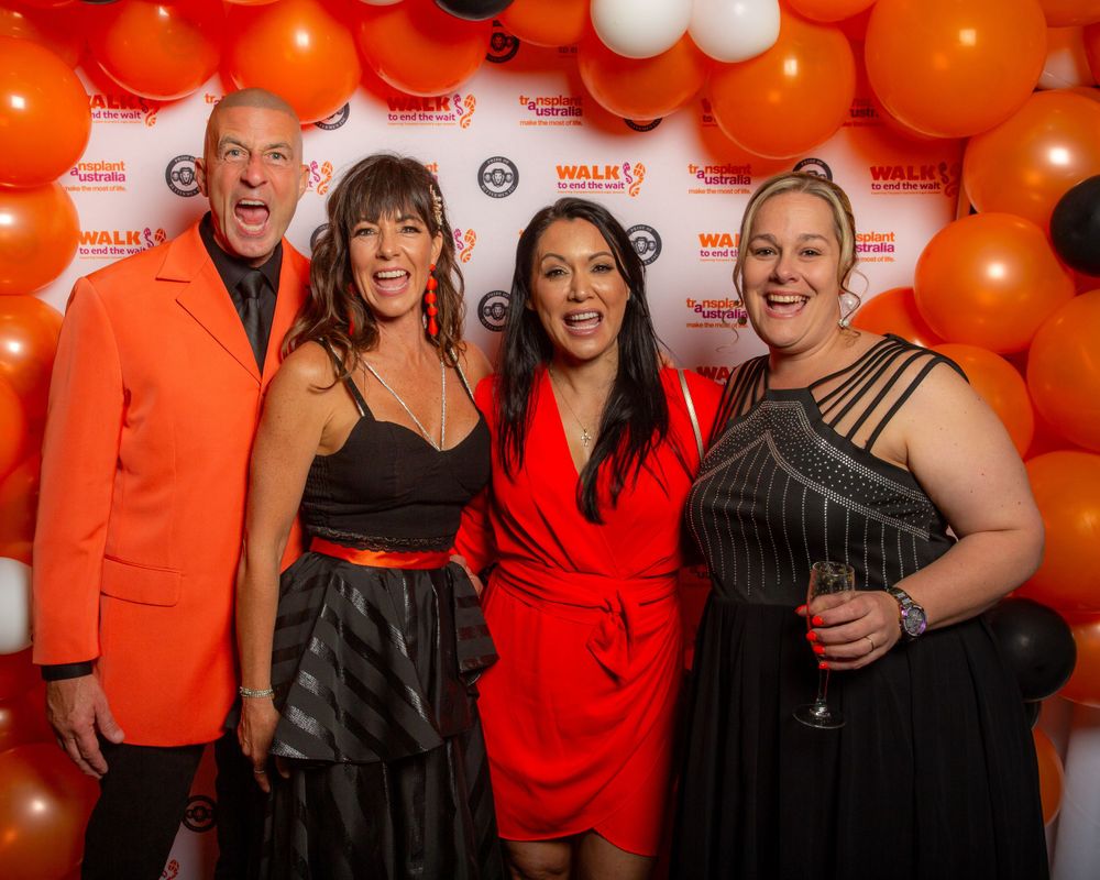 Four people pose at an event against an orange balloon arch backdrop wearing formal attire in red and black.