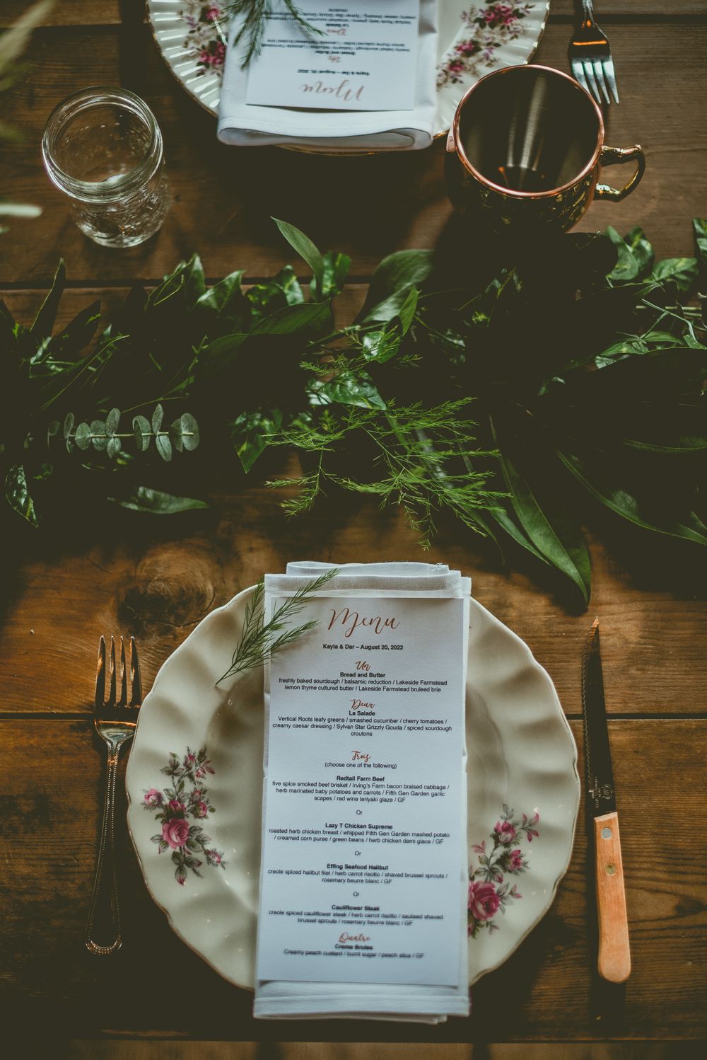 Elegant wedding table setting with floral china plate, menu card, and green foliage on wooden surface.