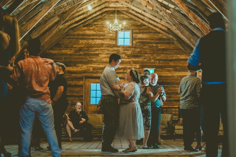 Couples dance together in a rustic wooden barn with exposed beams and small windows during a wedding celebration.