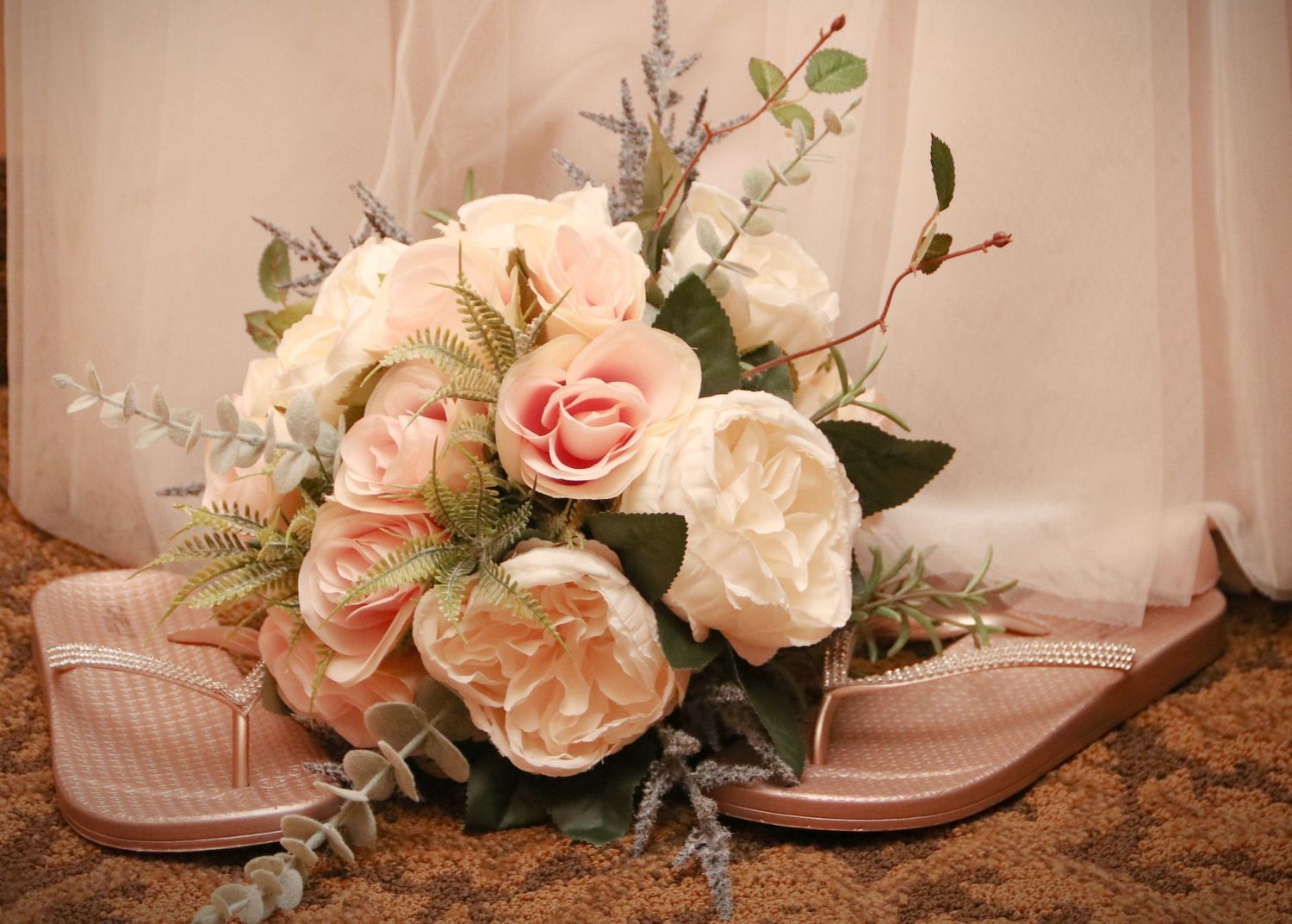 Pink and cream rose bouquet arranged with neutral sandals on brown surface.