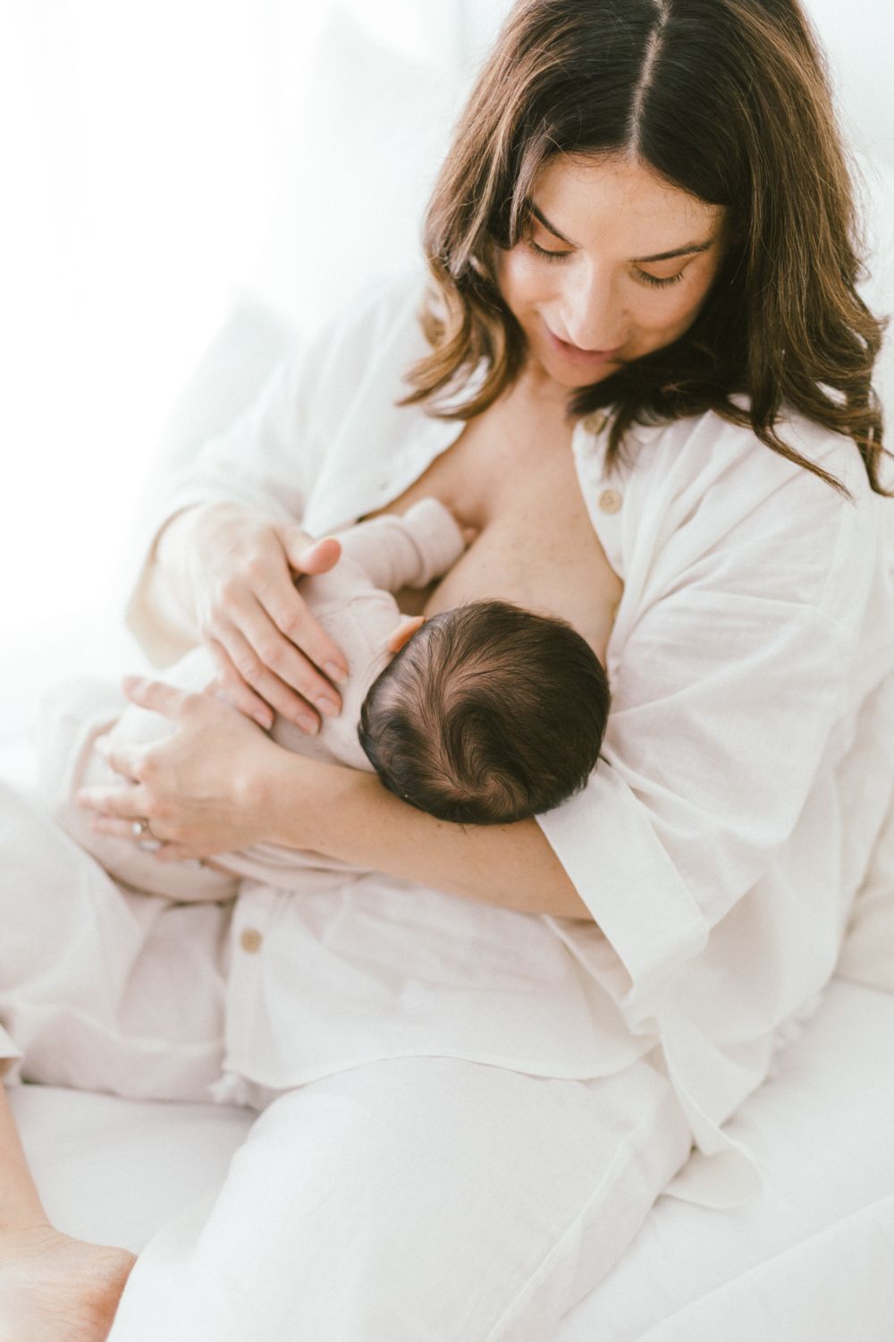 Intimate breastfeeding moment captured in soft natural light with white clothing.