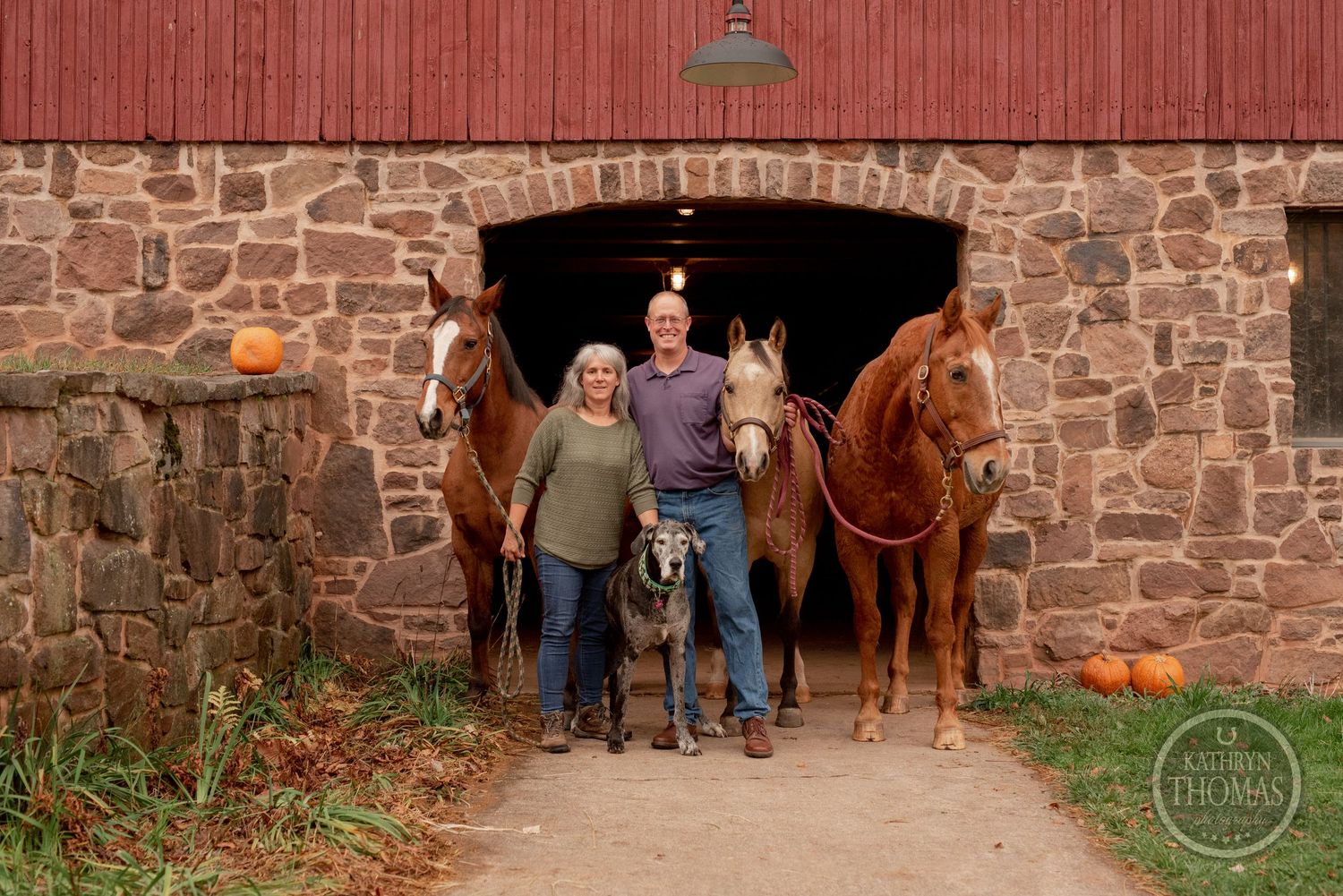 Two horses and their handlers stand in the stone archway of a red barn with pumpkins nearby.