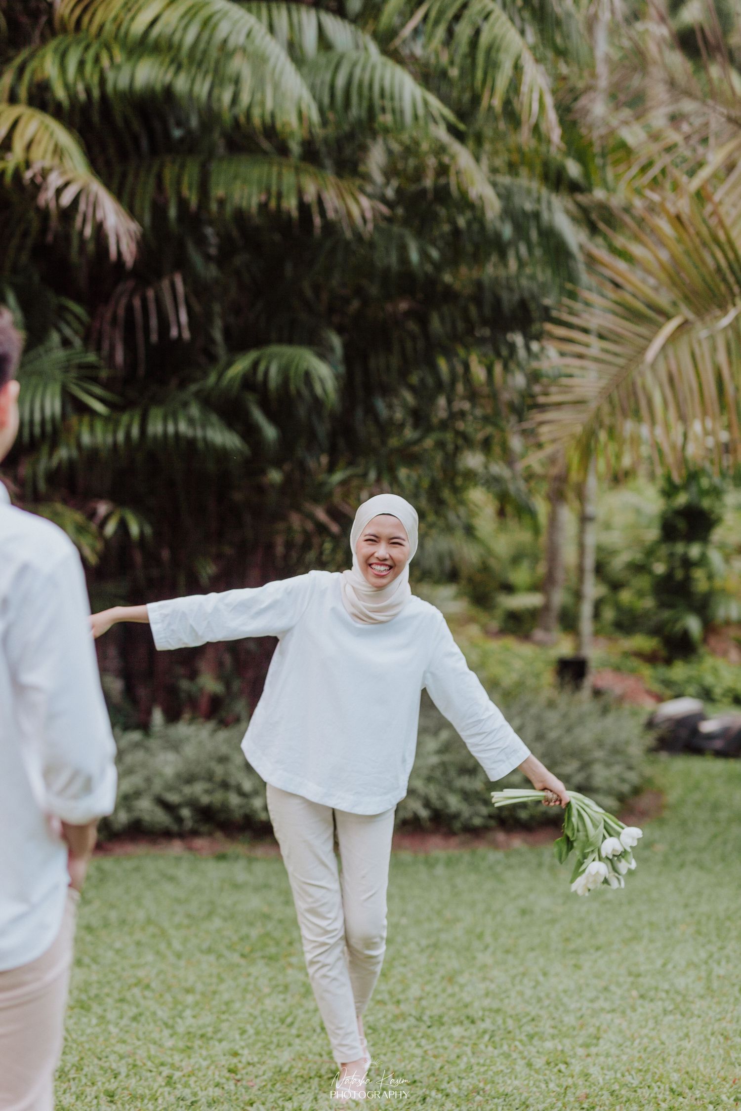 A person in all white clothing and hijab dances joyfully in a garden with palm trees.