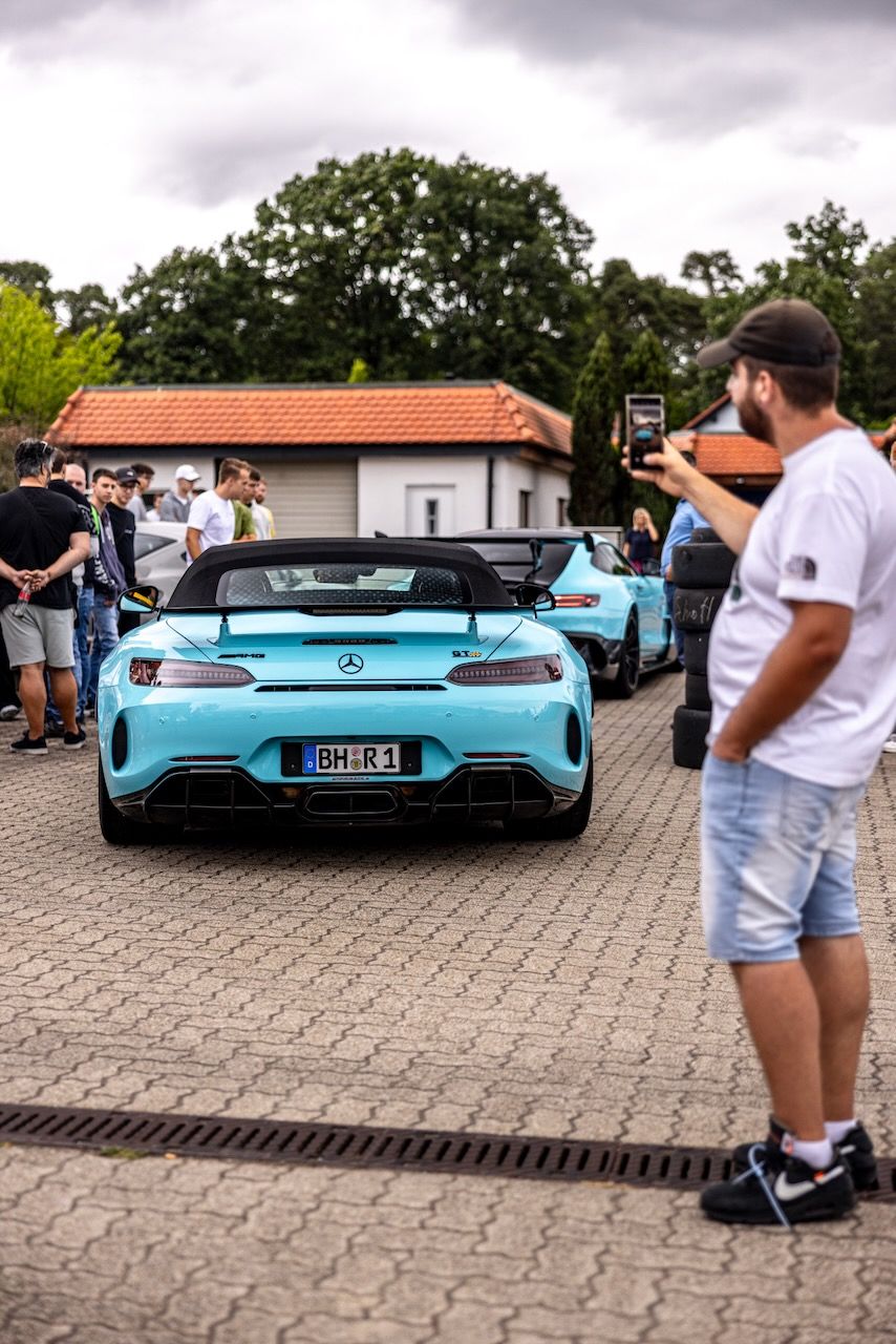 Ein hellblauer Mercedes AMG GT-R Roadster verlässt einen Parkplatz während er mit einem Smartphone gefilmt wird.
