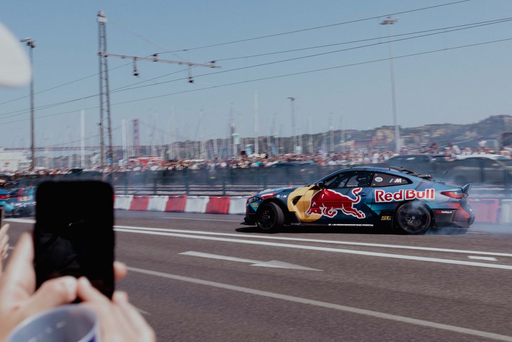 Red Bull branded BMW performing a drift demonstration on a street circuit with crowd recording on phones.
