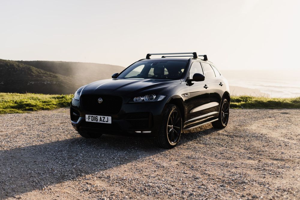 Black Jaguar F-Pace SUV parked on gravel road against coastal sunset landscape.