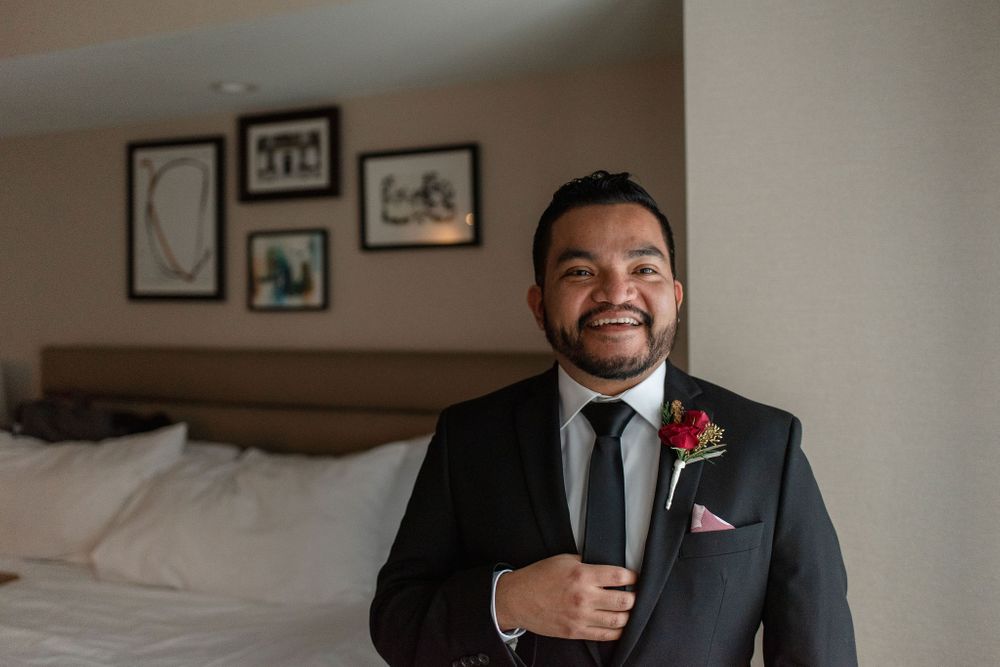 A groom in formal attire with red boutonniere smiles in front of framed wall art.