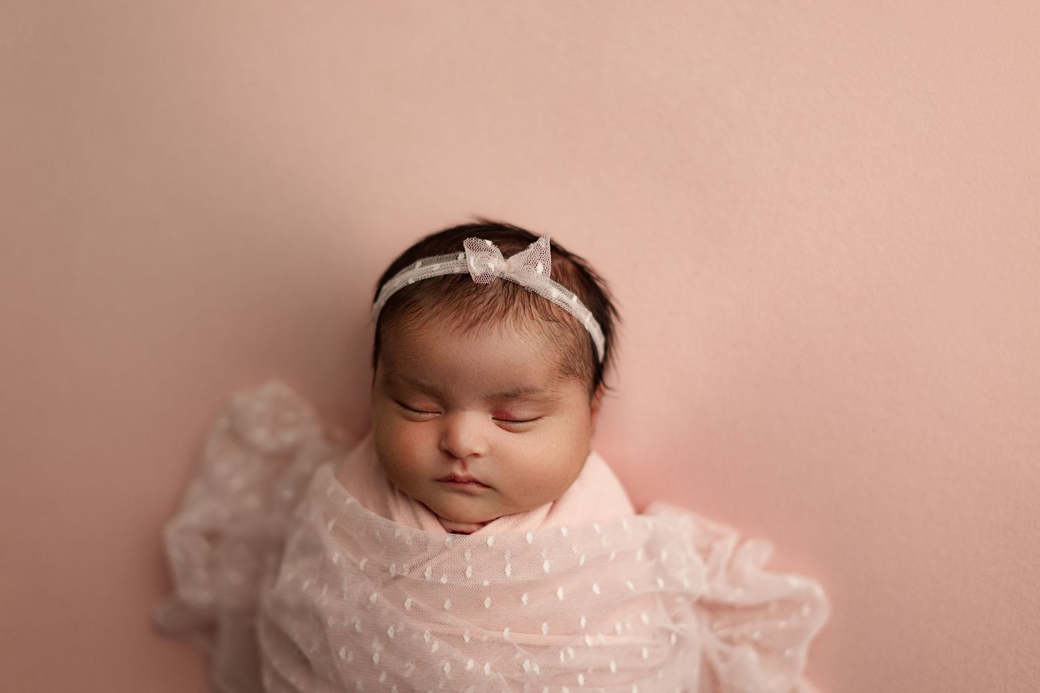 Sleeping newborn in delicate white lace dress with bow headband against pink backdrop.