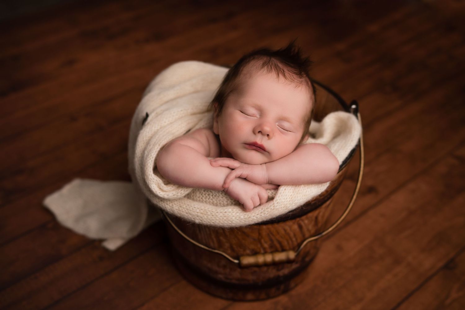 Sleeping newborn baby nestled in a brown basket with white fabric on wooden floor.