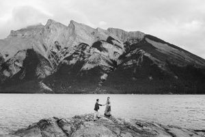 Black and white landscape of two figures standing on rocks beside a mountain lake.