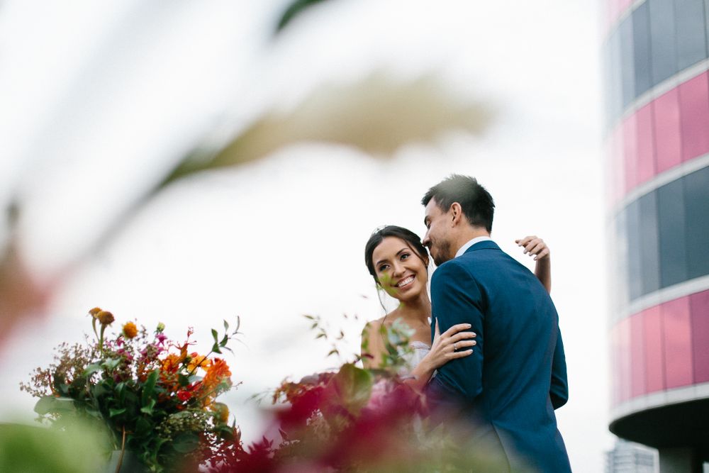A romantic moment between newlyweds surrounded by colorful flowers with urban architecture in background.