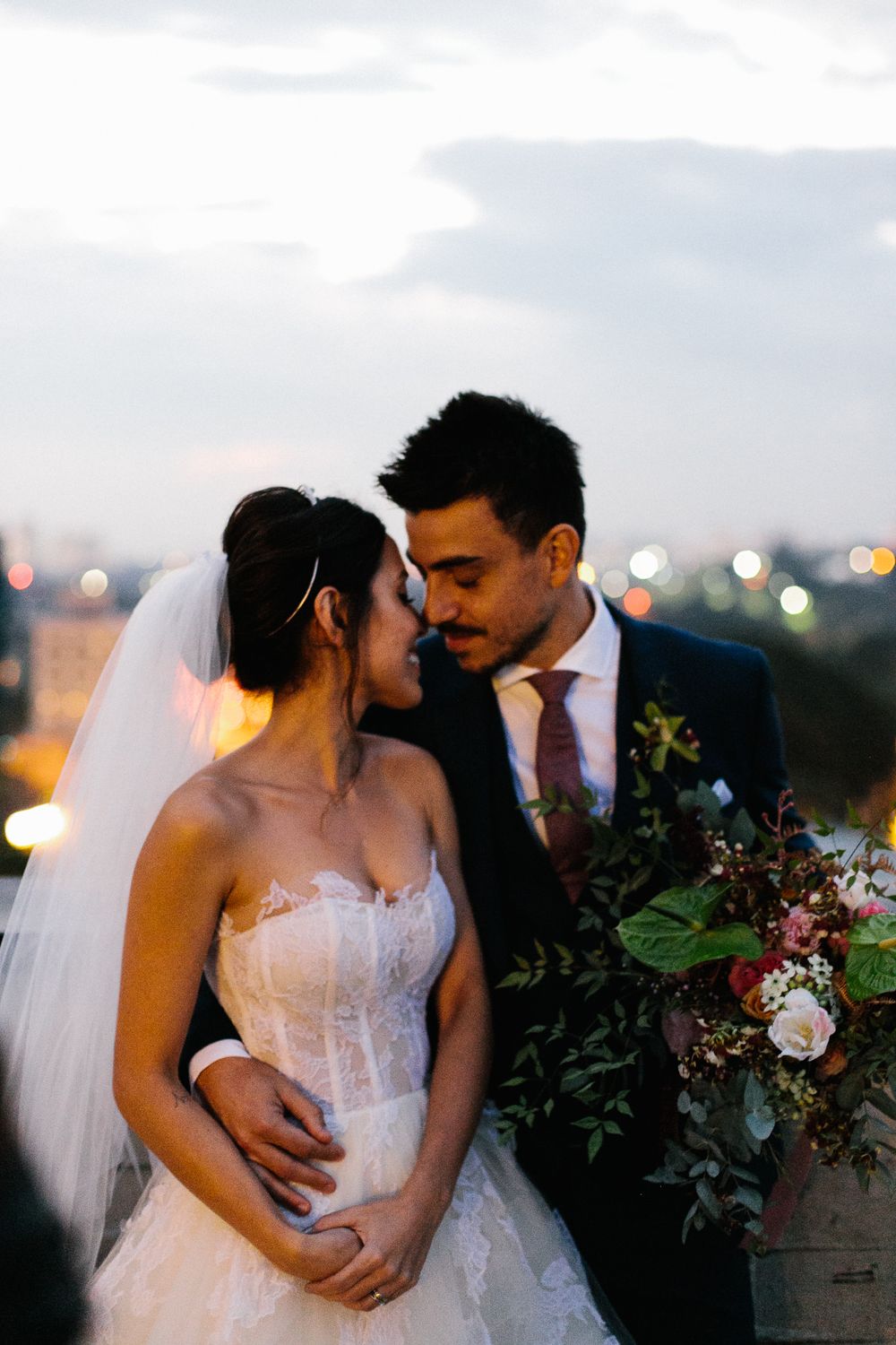 Romantic sunset portrait of newlyweds with city lights in background and beautiful floral arrangements.