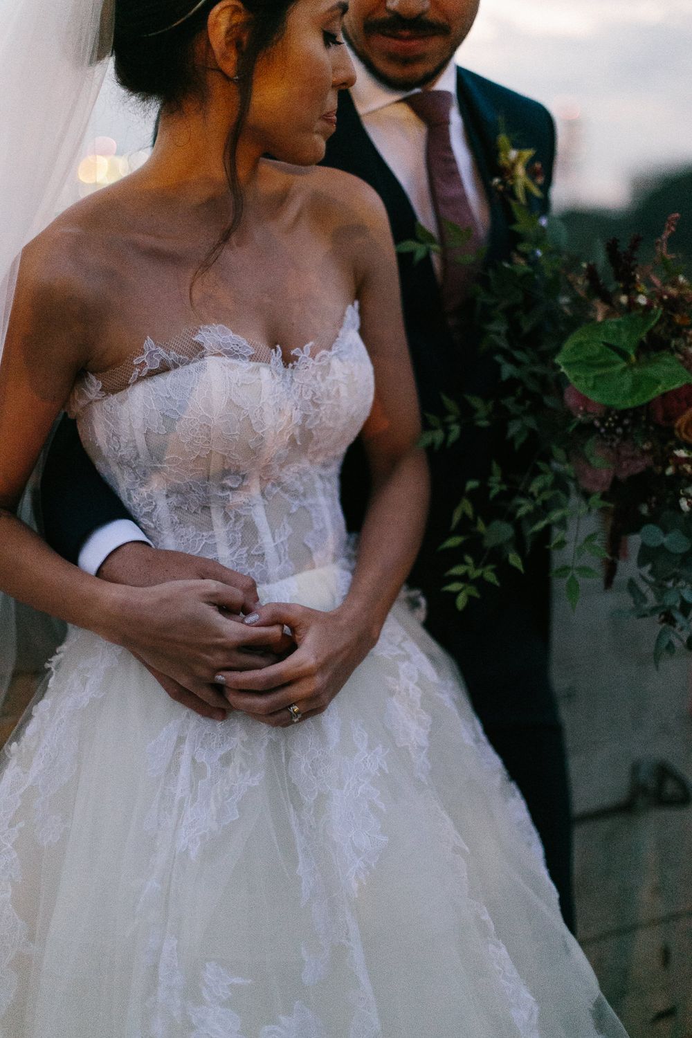 Bride in a stunning lace corset wedding dress embraced by her groom among greenery and florals.