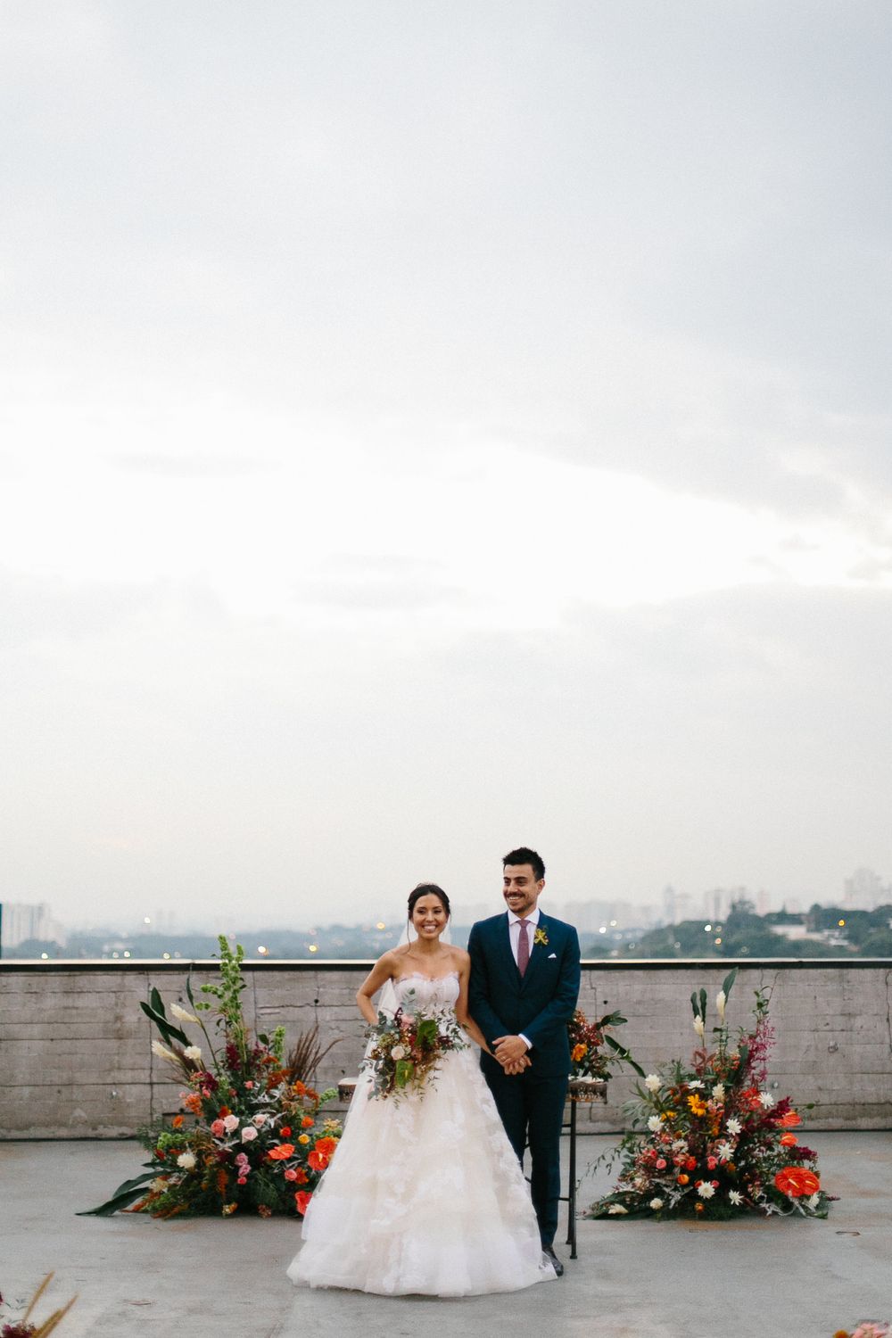 A wedding portrait of a couple standing between elegant floral arrangements on a rooftop venue.