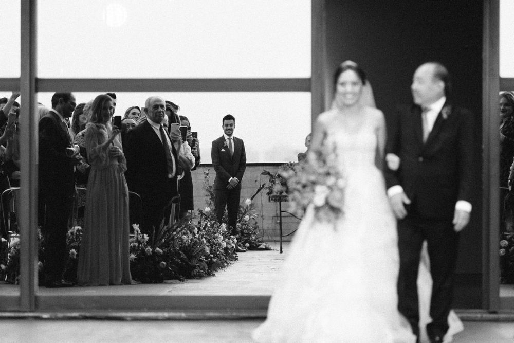Black and white wedding ceremony photo showing guests gathered in front of large windows.