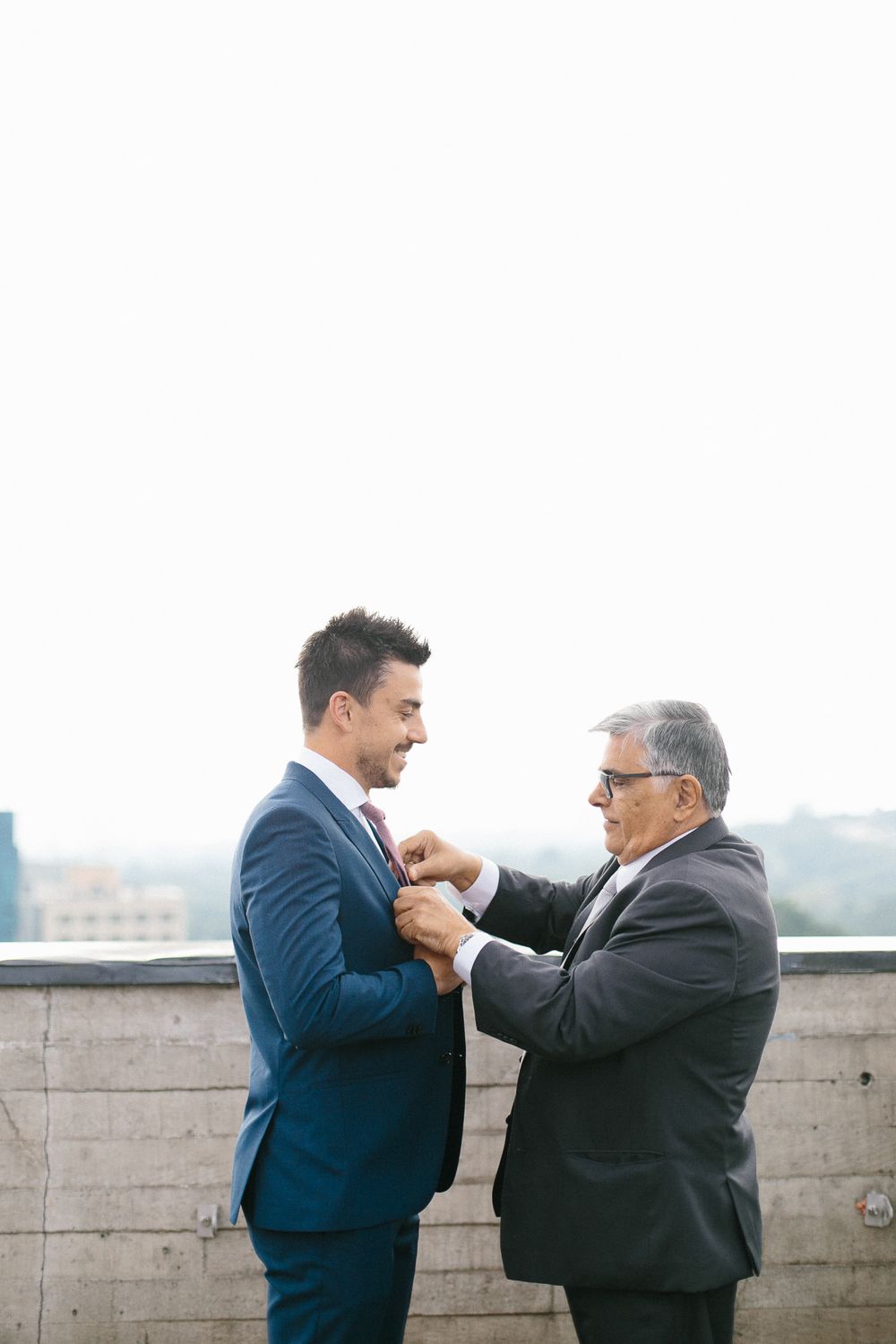 An older man helps adjust the younger man's suit jacket on a rooftop setting.