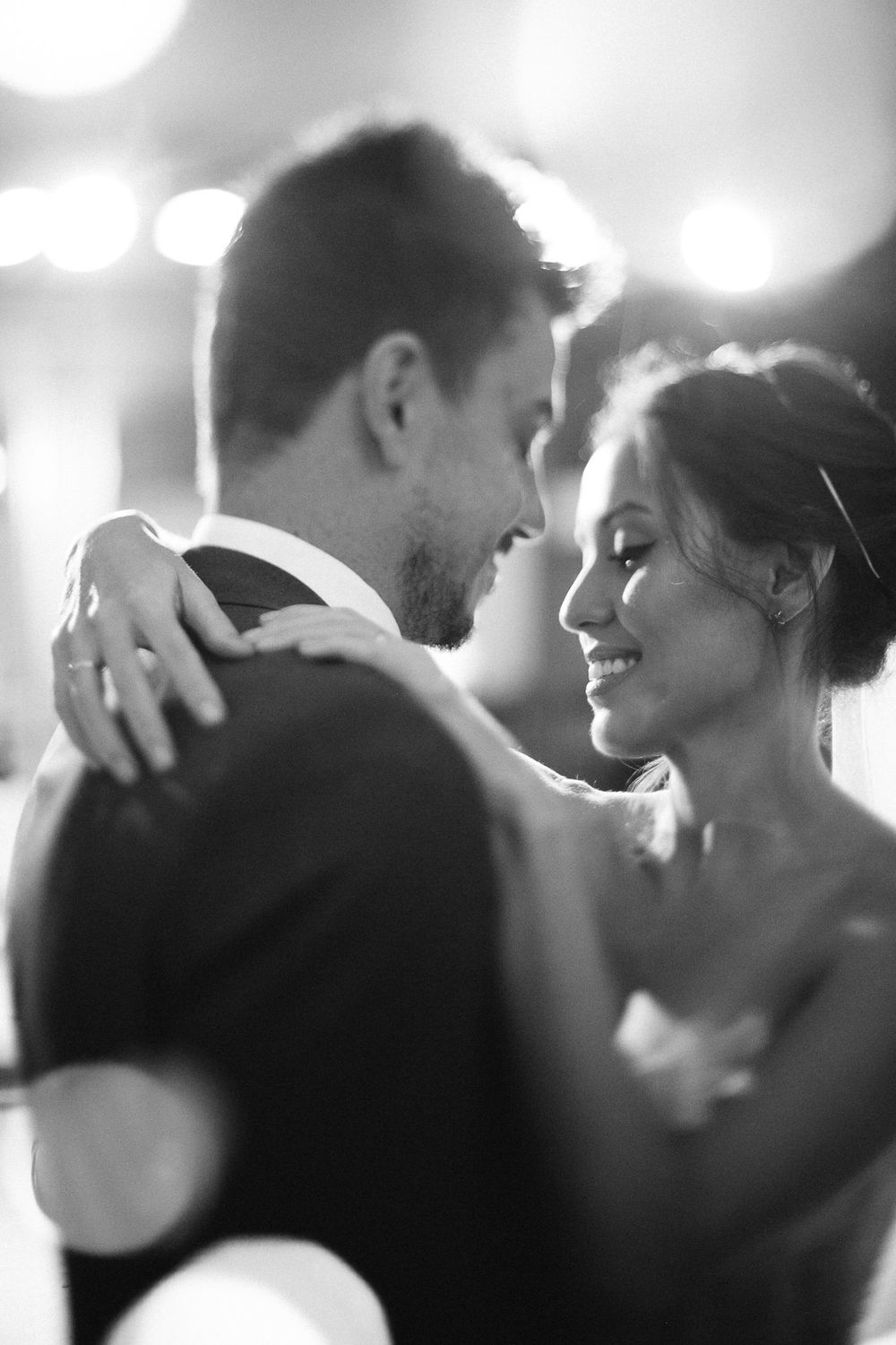 A tender moment captured in black and white as newlyweds share their first dance with soft bokeh lights in the background.