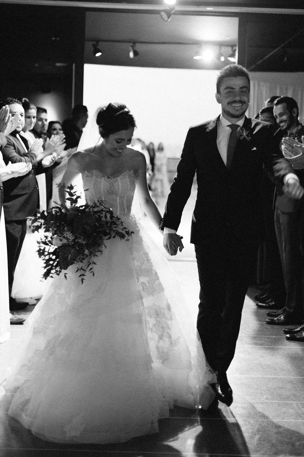 Newlyweds joyfully exit their ceremony while guests line the aisle in a black and white photo.