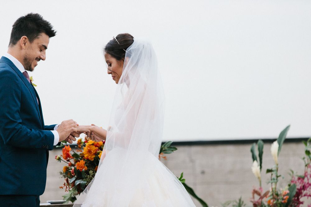 Couple exchanges rings during wedding ceremony against bright sky backdrop.