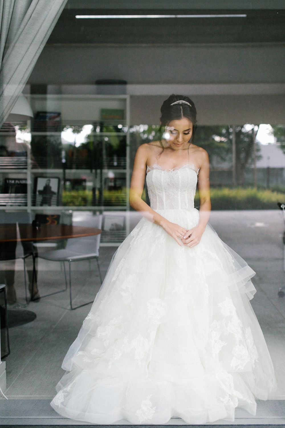 A bride in a classic white ball gown stands elegantly in front of reflective glass windows.