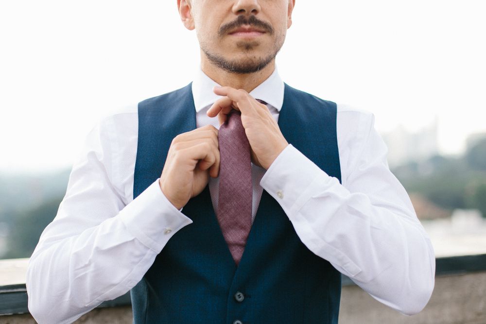Groom adjusting a burgundy tie while wearing a navy blue vest and white dress shirt.