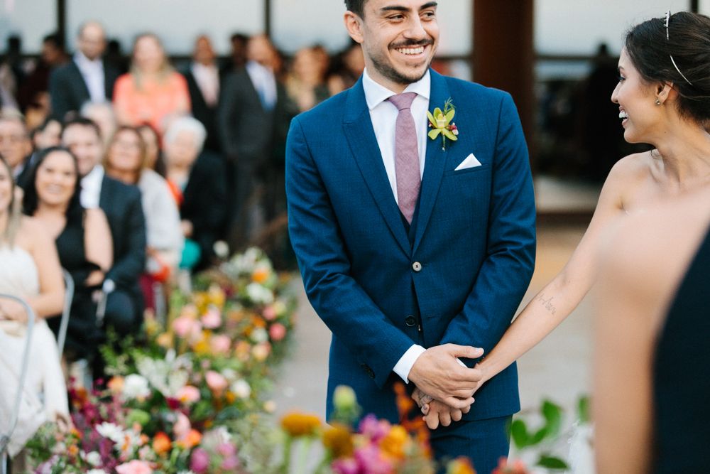 Newlyweds hold hands at altar surrounded by colorful floral arrangements during outdoor ceremony.