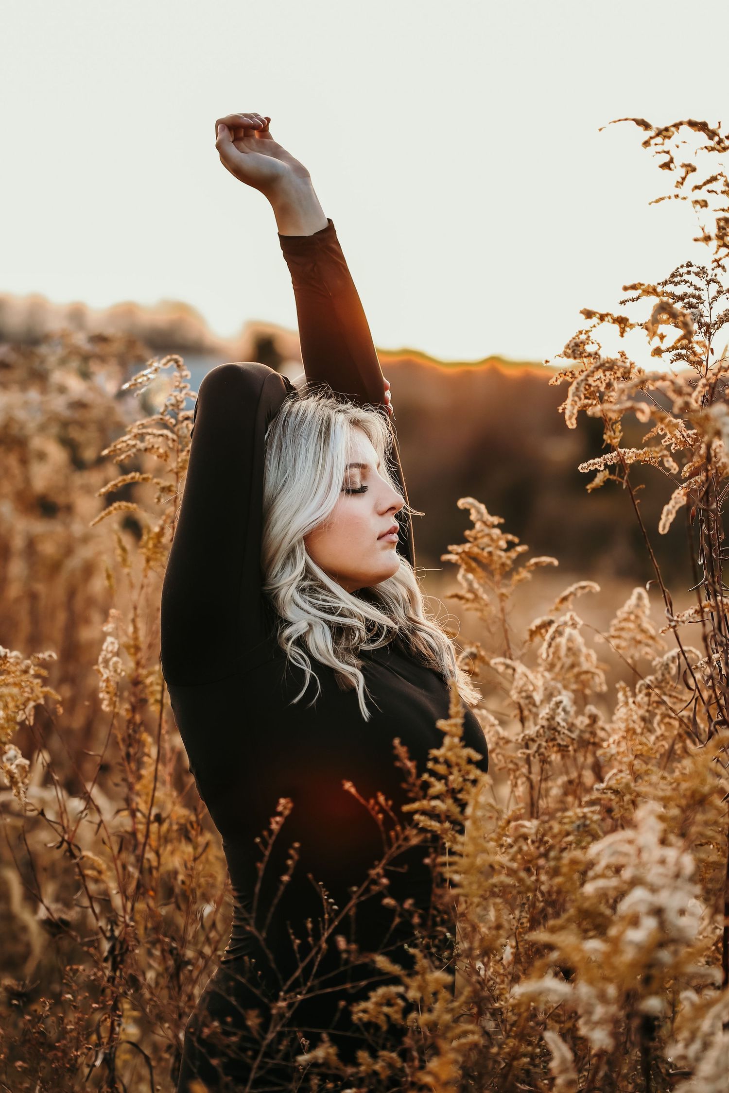 Woman in black stretches arms up during golden sunset in field of tall grass.