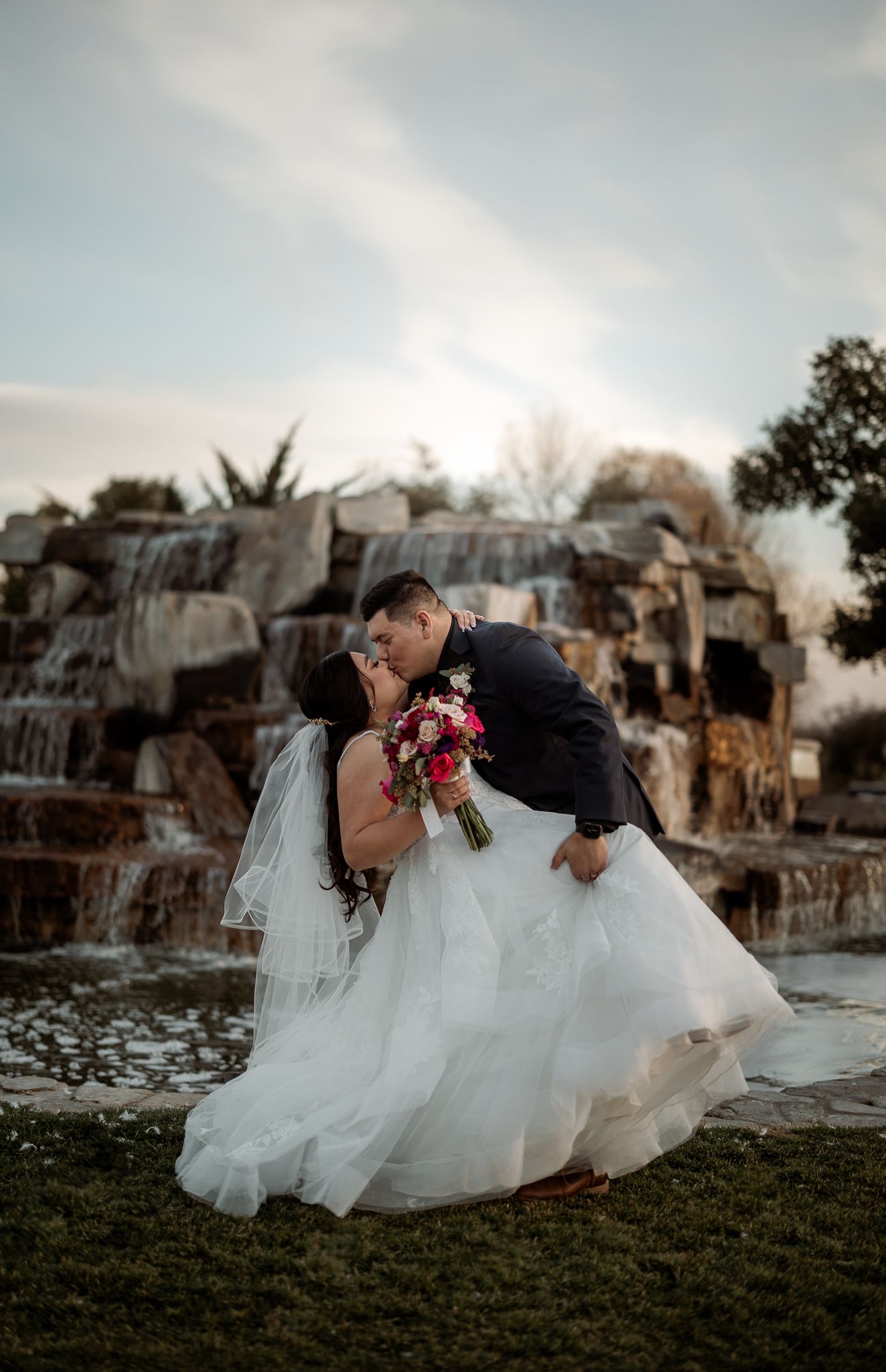 Romantic wedding couple sharing a kiss by a waterfall backdrop at sunset.