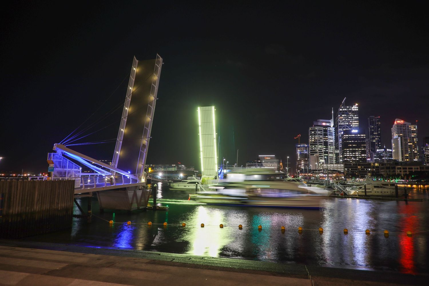 A raised drawbridge glowing with neon green and blue lights, casting colourful reflections across the calm waters of the harbour at night.