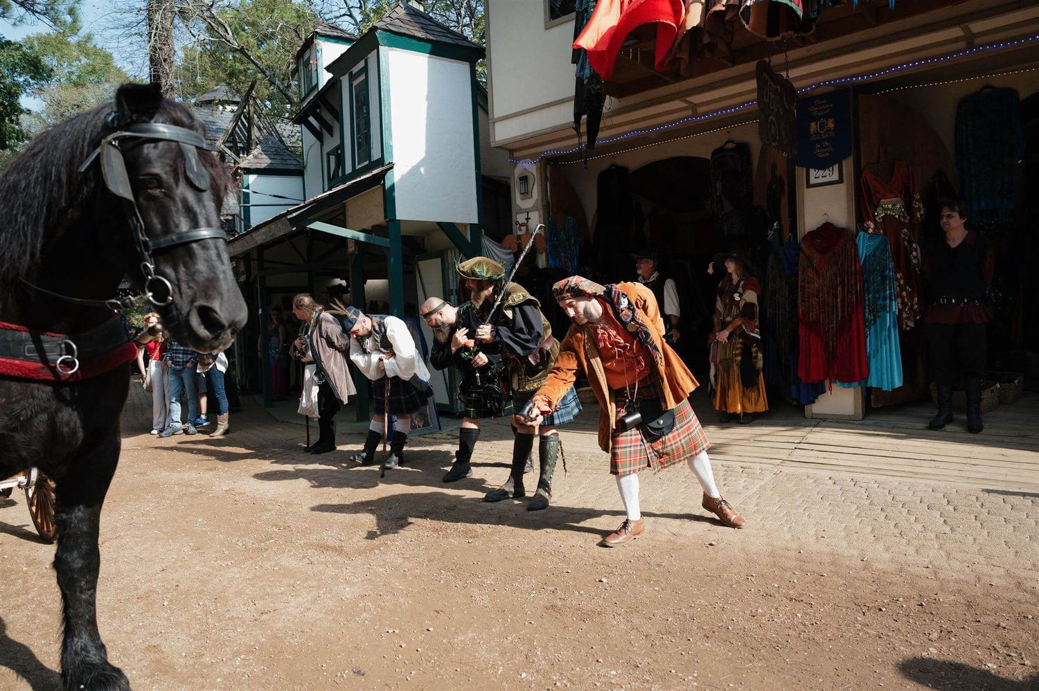 A black horse stands at a Renaissance fair while costumed performers interact in the background.