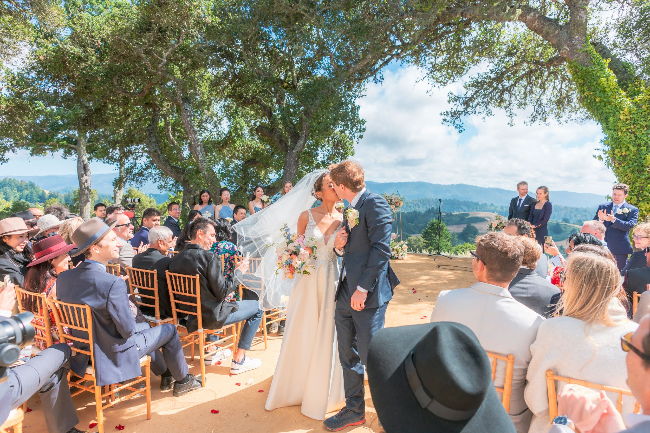 Bride and groom sharing a kiss at an outdoor wedding ceremony under oak trees.