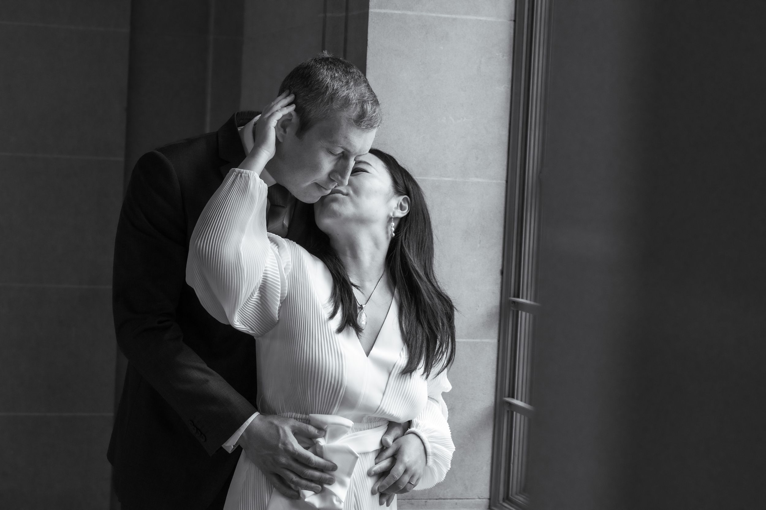 Black and white photo of a couple sharing a romantic embrace near a window.