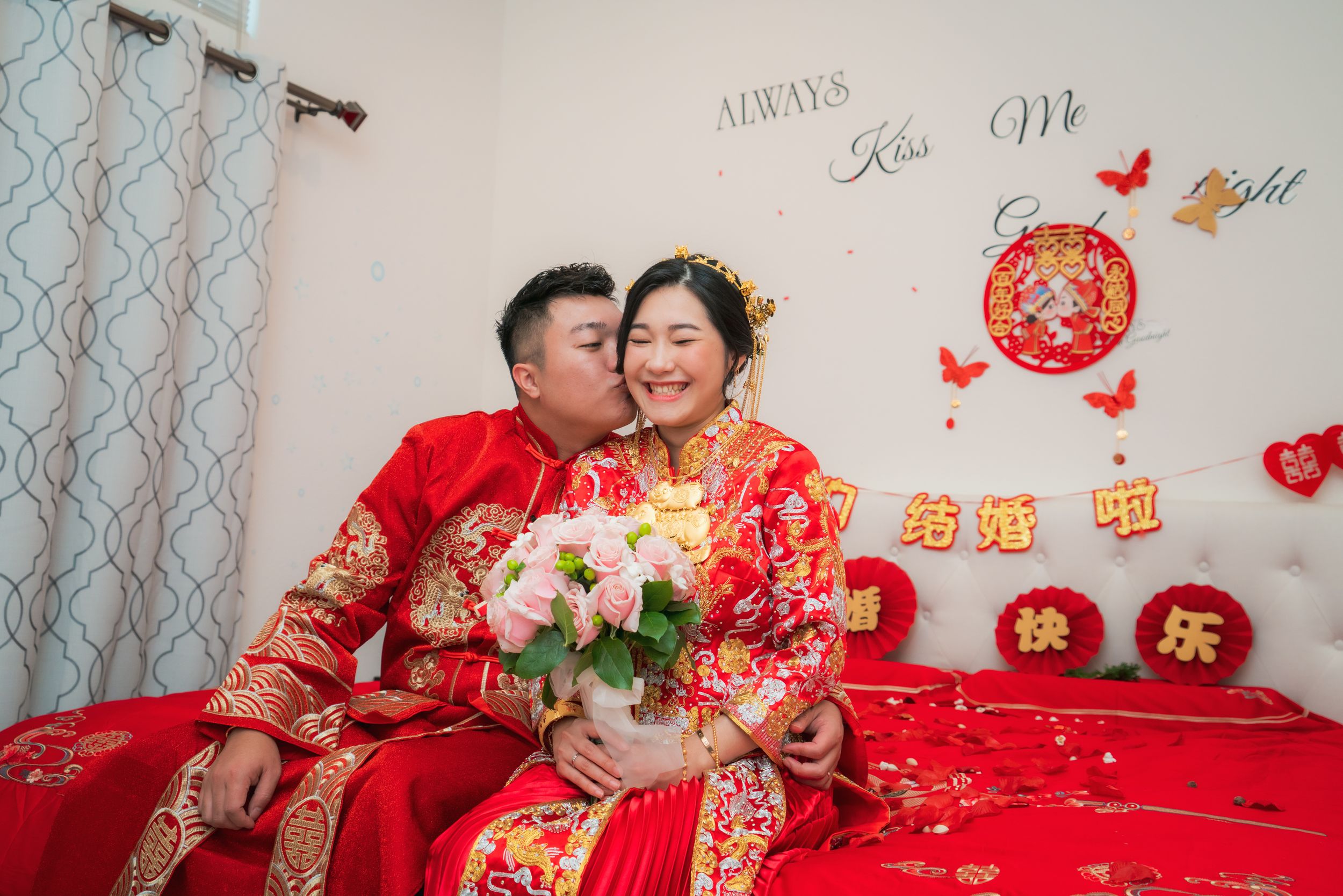 Chinese couple in traditional red qipao wedding attire sharing a kiss on a decorated bed.