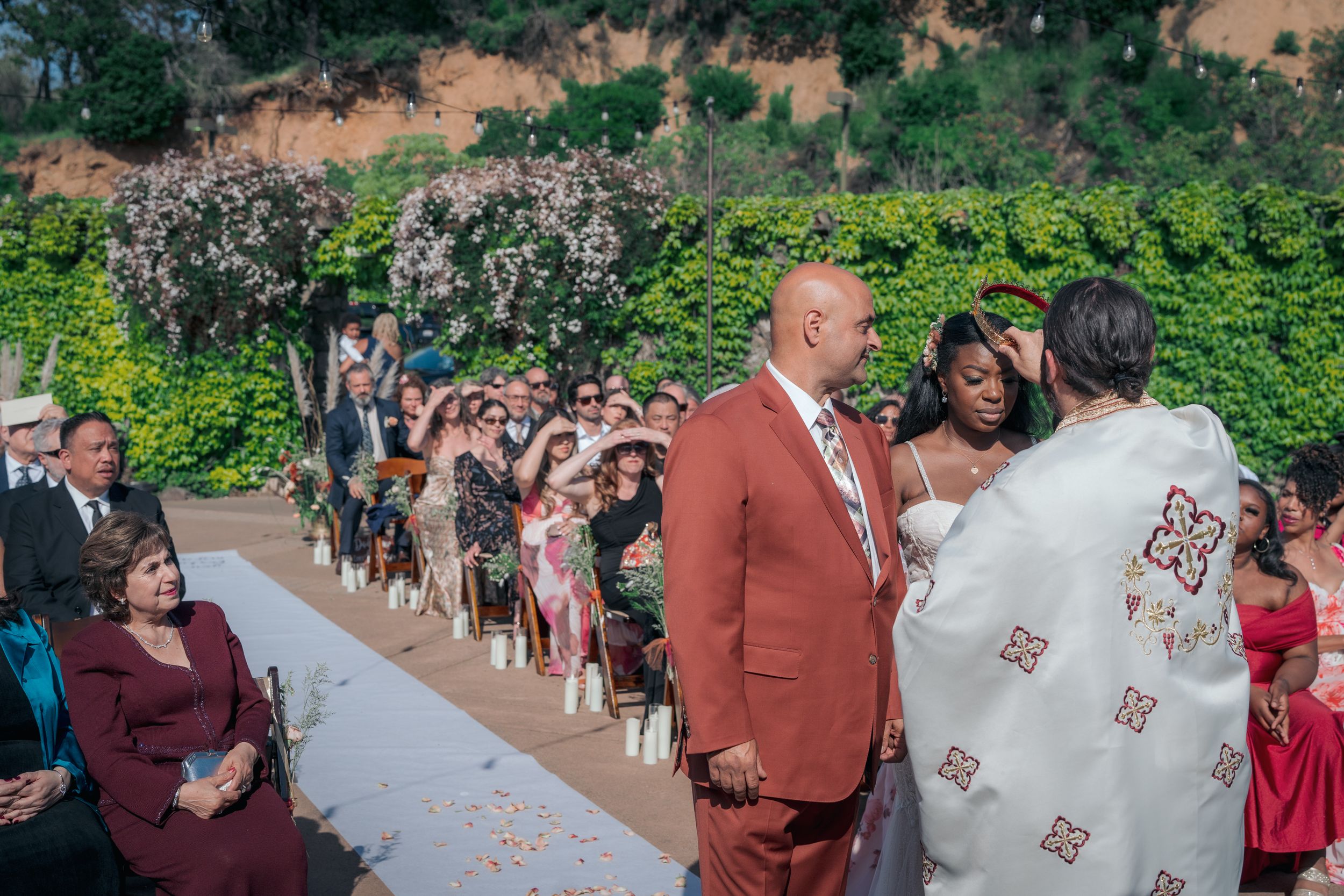 Groom in terracotta suit awaiting bride during an outdoor wedding ceremony with guests seated.