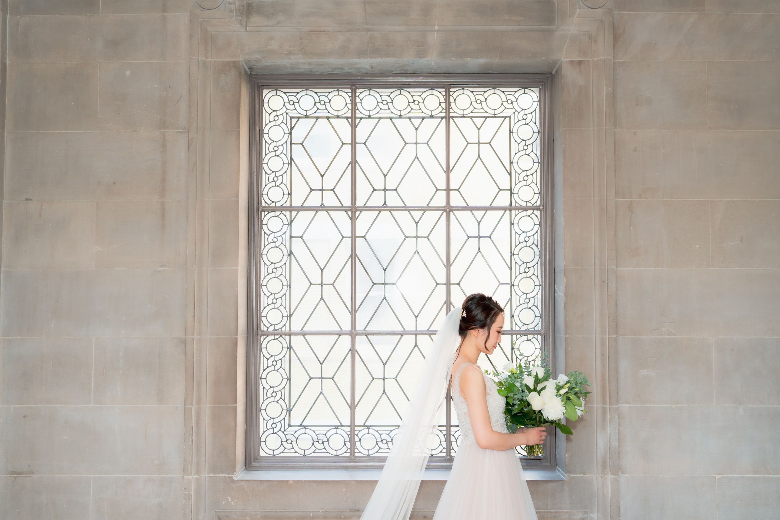 Elegant bride in white gown holding bouquet standing before ornate window in San Francisco City Hall.