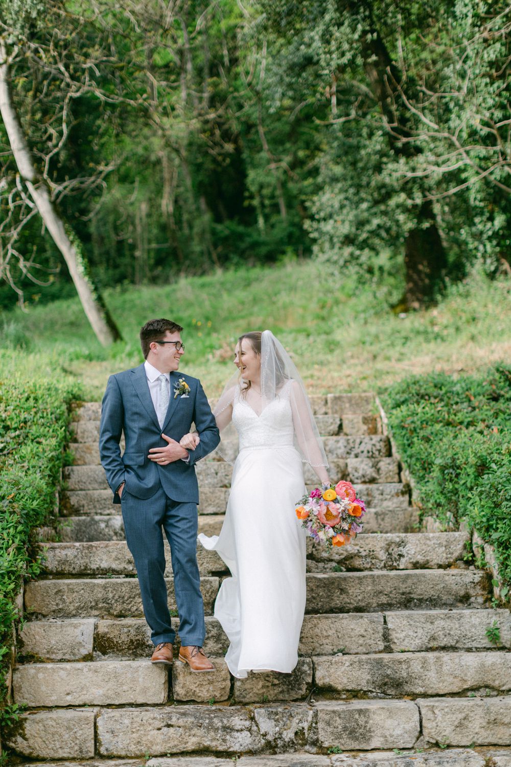 Le couple de mariés se tient sur des marches en pierre rustiques, entouré de verdure.