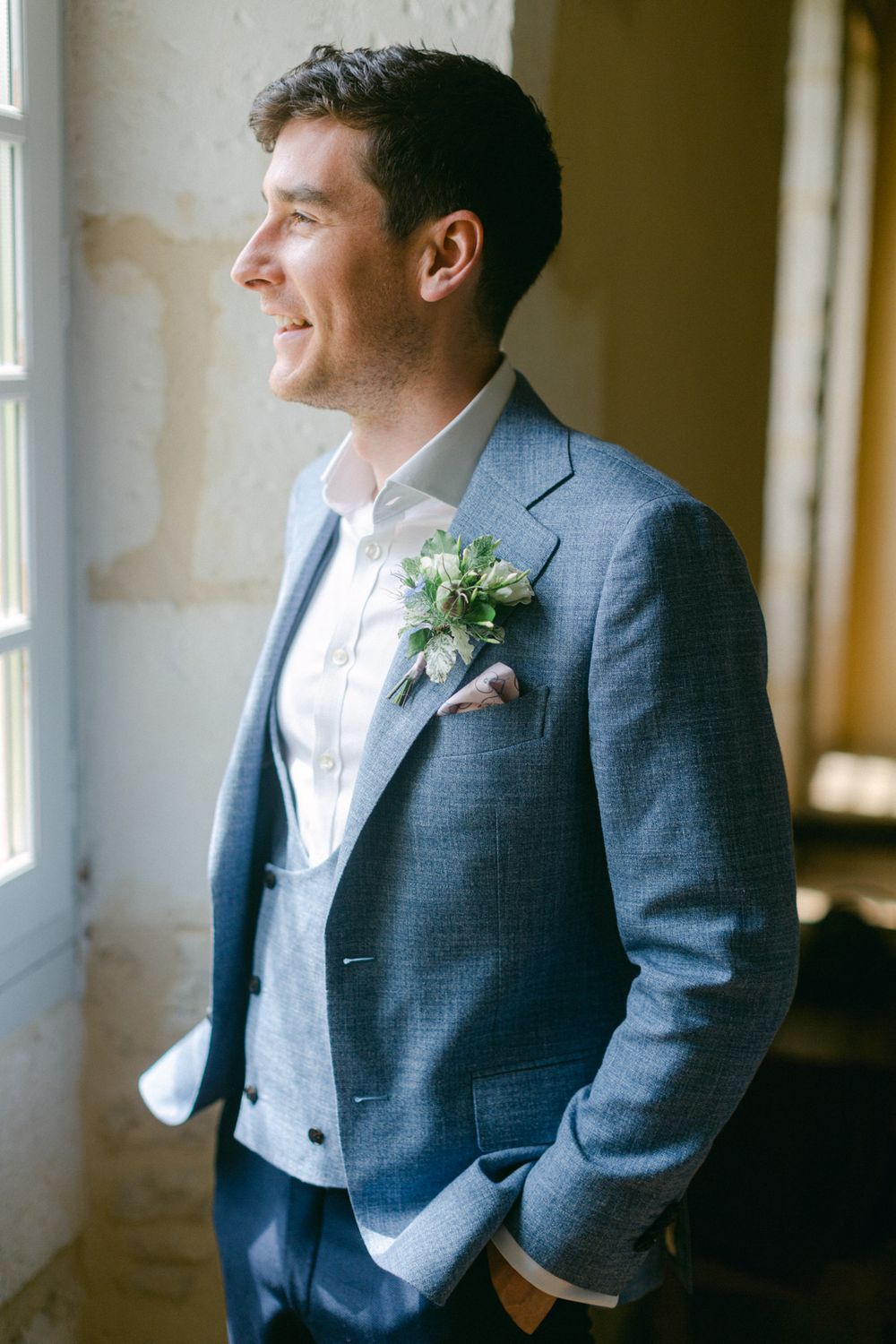 Le marié en costume bleu clair avec une boutonnière sourit près de la fenêtre, baigné de lumière naturelle.