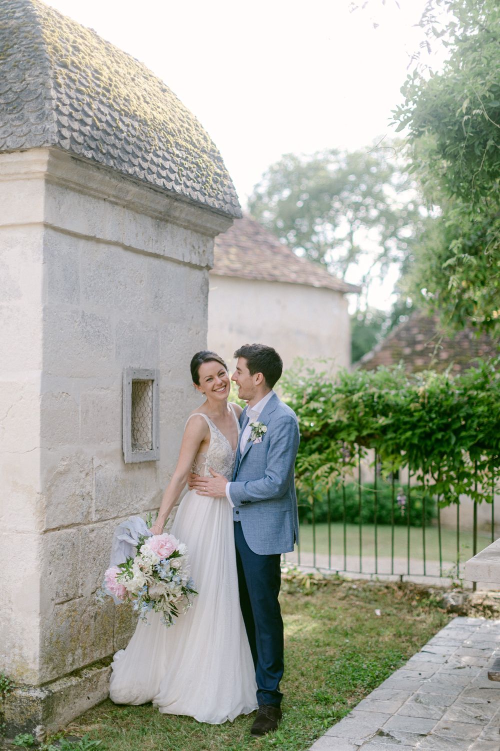 Le couple de mariés s’enlace près d’un bâtiment en pierre, avec un magnifique bouquet et une architecture classique en arrière-plan.