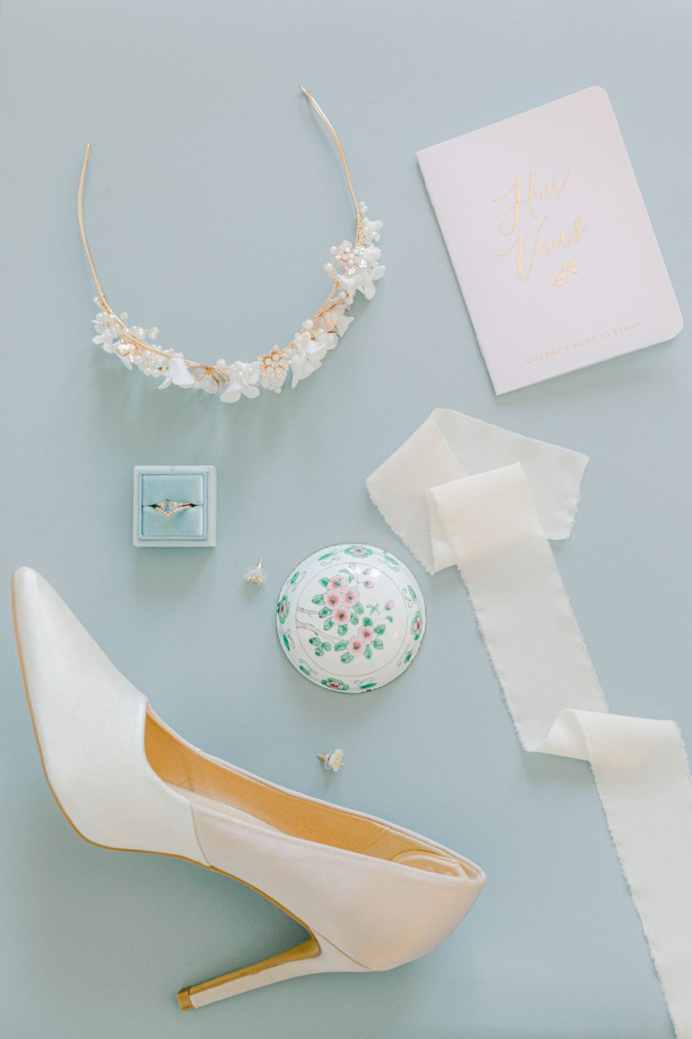 Accessoires de mariage disposés à plat avec une chaussure blanche à talon, une couronne de fleurs et des bijoux sur un fond bleu.