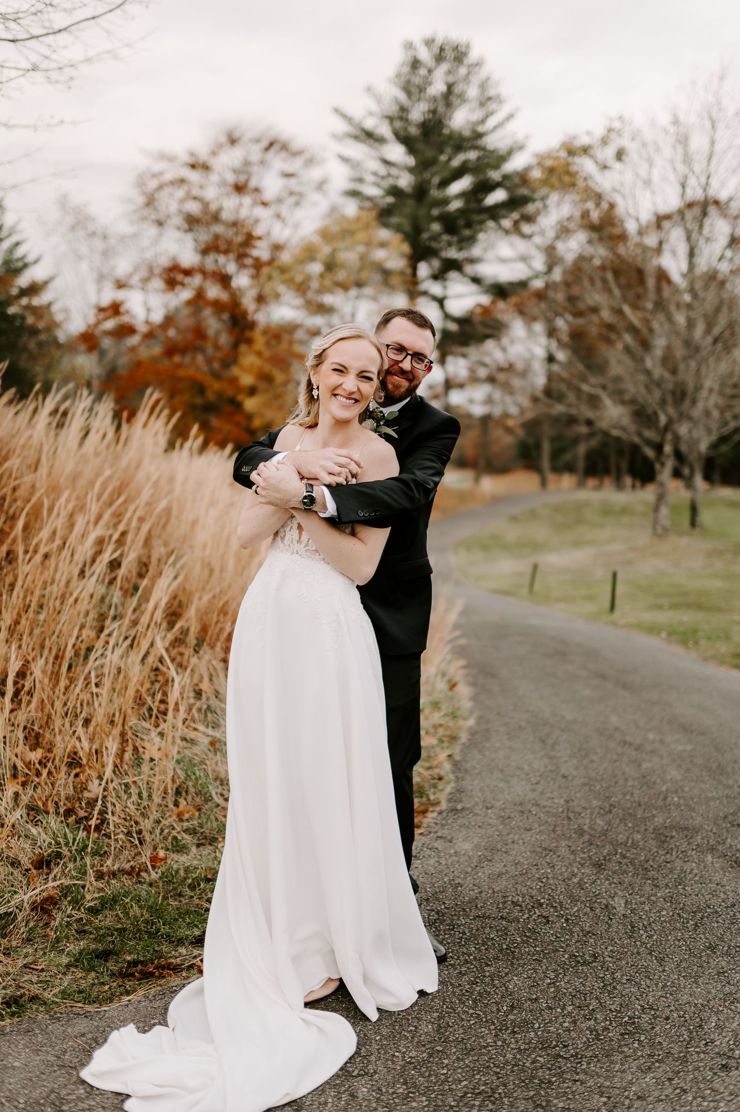 A romantic wedding photo of a couple embracing on a path with autumn foliage and tall grasses in the background.
