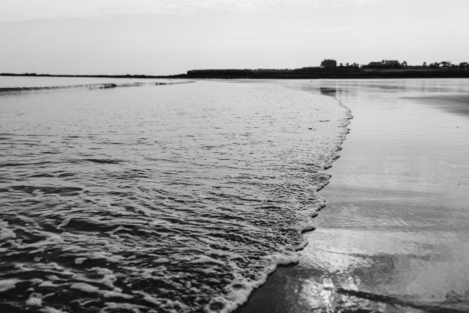 Waves rolling onto sandy beach with distant shoreline in black and white.