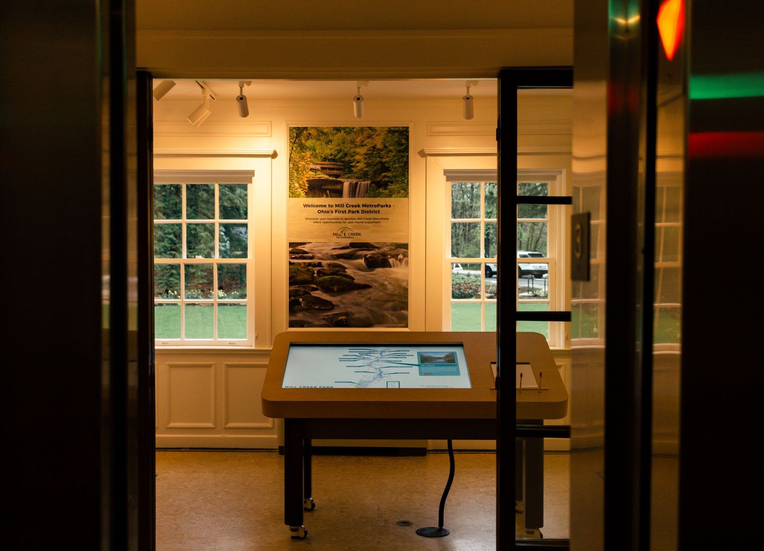 Museum room with digital display table and framed artwork visible through doorway.