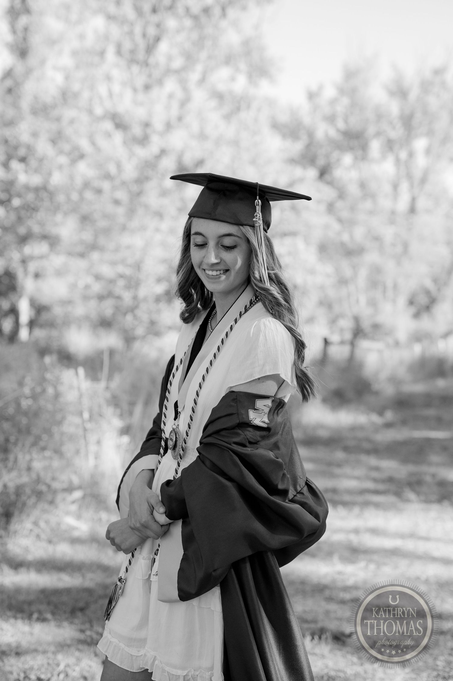 Black and white portrait of graduate in traditional cap and gown surrounded by spring blossoms.