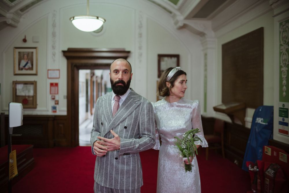 Wedding couple stands in ornate hallway with arched ceilings and red carpet.
