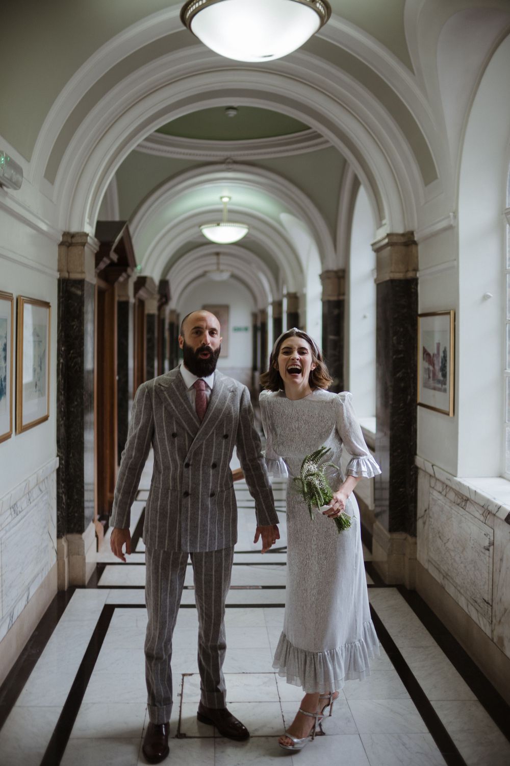 Couple walks through elegant arched hallway in vintage-inspired wedding attire.