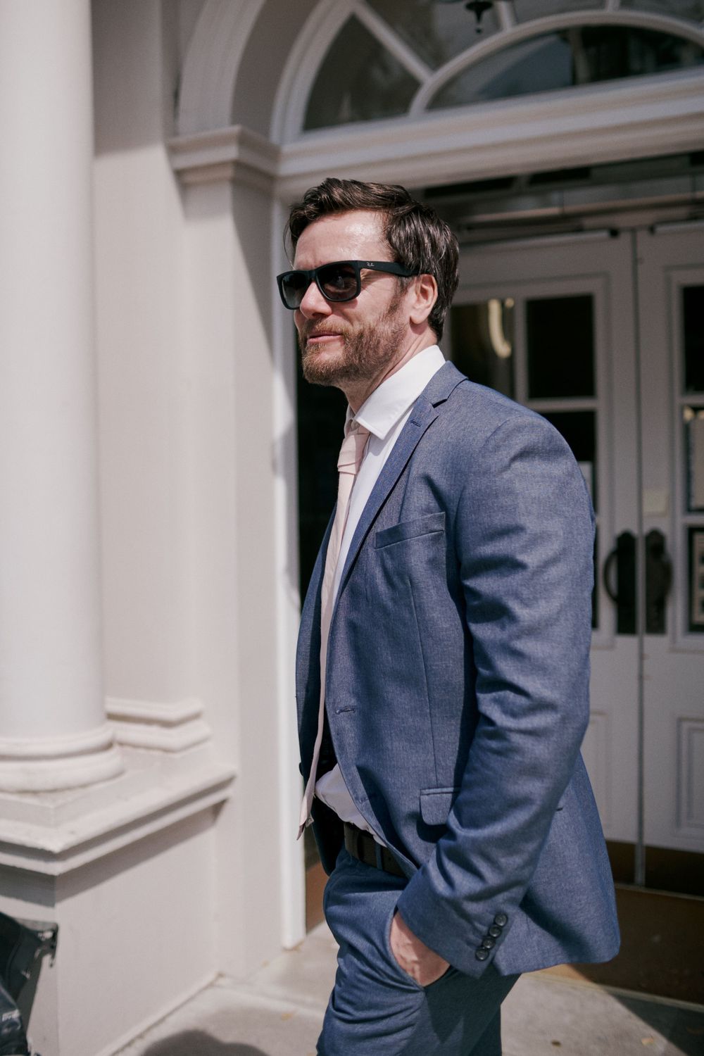 A stylish man in a blue suit and sunglasses poses outside a building with white columns.