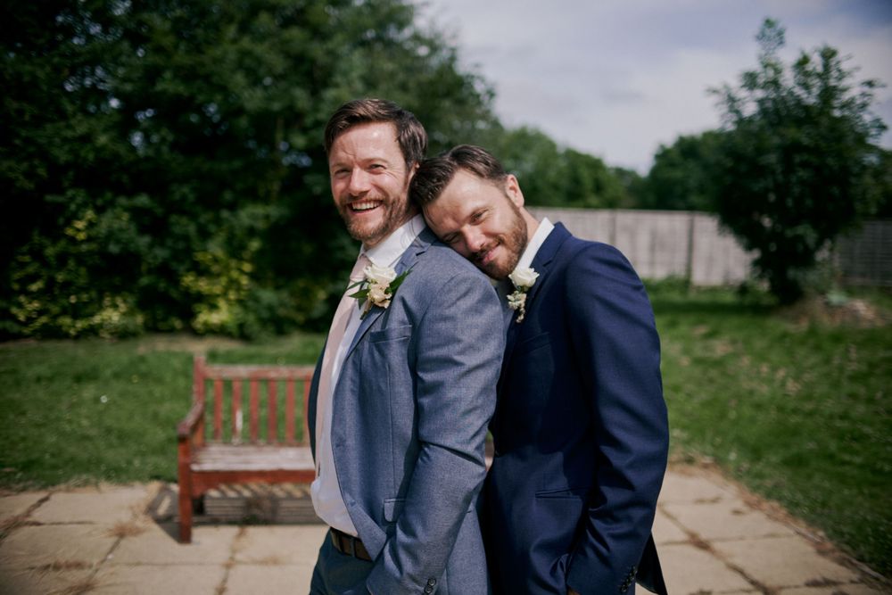 Two people in suits share a playful moment in a garden setting.