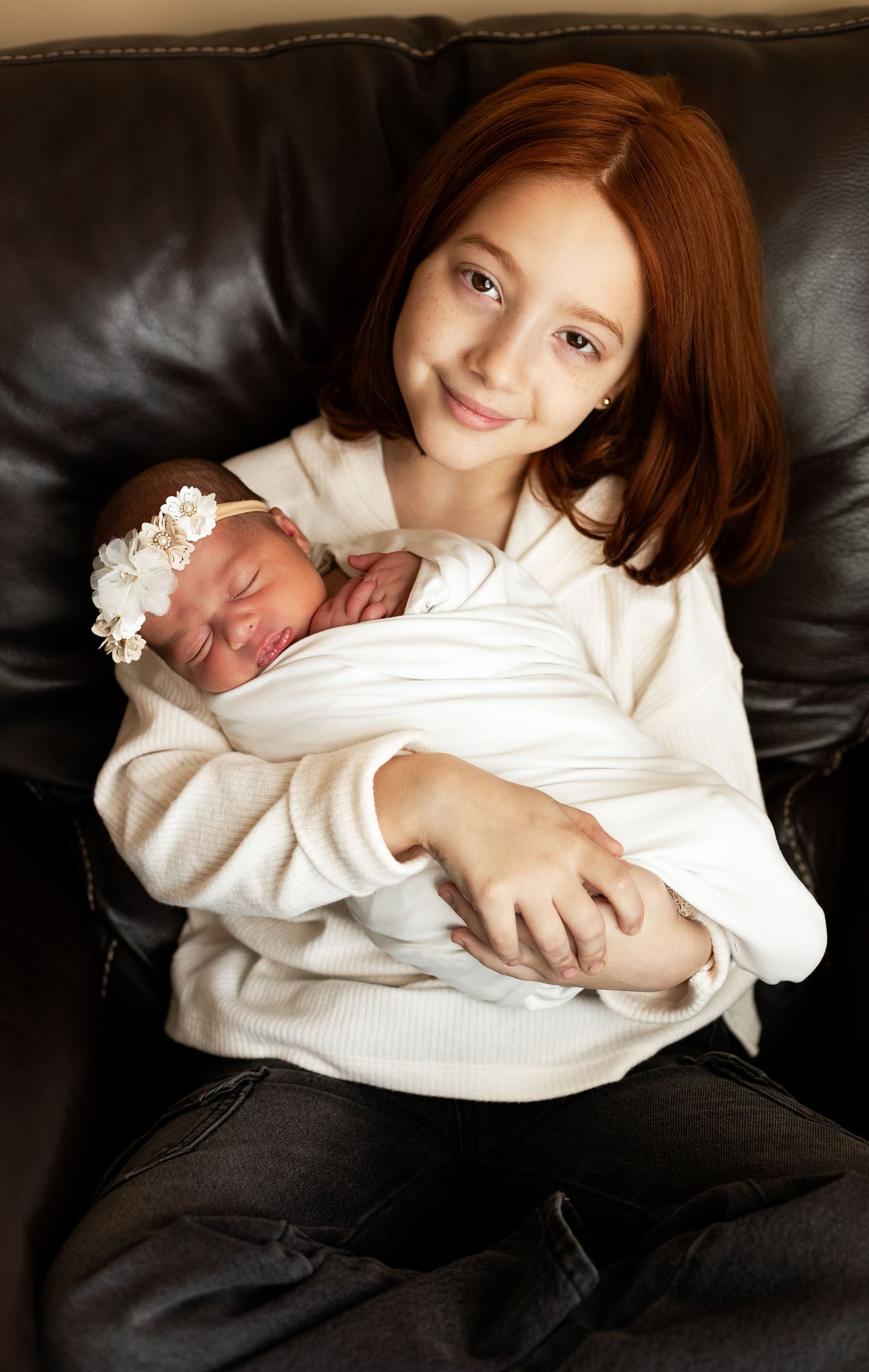 Tender moment between siblings as older child holds newborn on black leather couch.